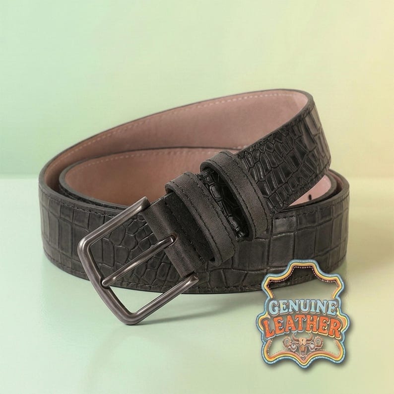 Pu&ograve; includere: Una cintura in pelle grigio scuro con un motivo strutturato a forma di coccodrillo. La cintura ha una fibbia color argento e un interno rosa chiaro. &Egrave; visibile un emblema "GENUINE LEATHER".