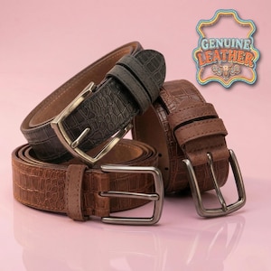 Könnte beinhalten: Drei Echtleder-Gürtel in Braun- und Grautönen mit silberfarbenen Schnallen. Die Gürtel haben ein strukturiertes, krokodilartiges Muster. Das Bild enthält ein dekoratives Schild mit der Aufschrift "GENUINE LEATHER".
