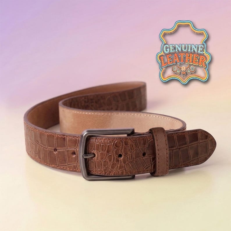 Pu&ograve; includere: Una cintura in pelle marrone con motivo a coccodrillo. La cintura ha una fibbia rettangolare in metallo scuro e un interno beige chiaro. L'immagine include un emblema "GENUINE LEATHER".
