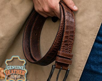 Può includere: Una cintura in pelle marrone con una texture a pelle di coccodrillo. La cintura ha una fibbia in metallo scuro ed è tenuta da una persona. L'immagine include un logo "GENUINE LEATHER".