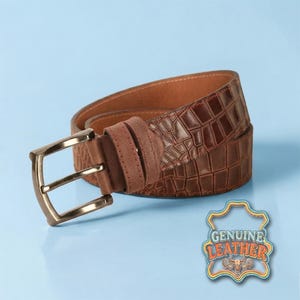 Pu&ograve; includere: Una cintura in pelle marrone con texture a coccodrillo e fibbia color oro. La cintura ha un passante in pelle scamosciata ed &egrave; etichettata "GENUINE LEATHER". La cintura &egrave; esposta su sfondo azzurro.