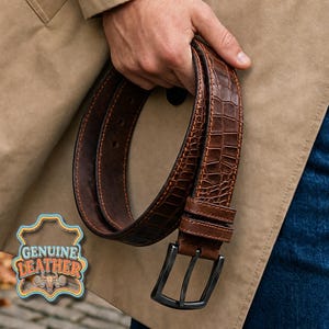 Pu&ograve; includere: Una cintura in pelle marrone con una texture a pelle di coccodrillo. La cintura ha una fibbia in metallo scuro ed &egrave; tenuta da una persona. L'immagine include un logo "GENUINE LEATHER".