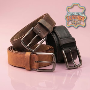 Puede incluir: Tres cinturones de cuero en tonos marrón y negro apilados sobre una superficie rosa. Cada cinturón tiene una hebilla de color plateado. La imagen incluye un gráfico que dice "GENUINE LEATHER".