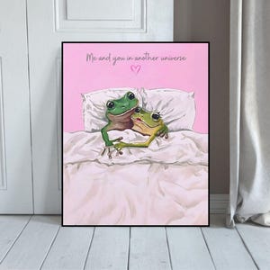 Peut inclure: Impression d'art encadrée représentant deux grenouilles de dessin animé au lit, s'embrassant sous une couette blanche. Le fond est rose, avec le texte "Me and you in another universe" et un cœur.