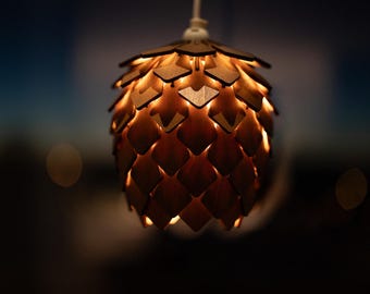 Puede incluir: Una lámpara colgante de madera con forma de piña. La lámpara está hecha de piezas de madera entrelazadas que crean una superficie texturizada. La luz brilla a través de las piezas de madera, creando un brillo cálido. Cuelga de un cable blanco.