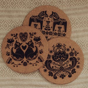 May include: Three round cork coasters with black folk art designs. One coaster features two horses, another has two foxes with a heart, and the third has a floral pattern. The coasters are arranged on a beige patterned surface.