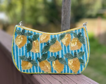 May include: A beaded clutch purse with a blue and white striped background. The purse is decorated with yellow lemons and green leaves. The purse has a yellow trim and a silver chain strap.