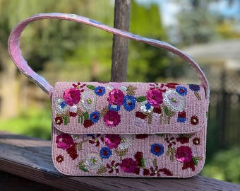 Könnte beinhalten: Eine hellrosa Perlenhandtasche mit floralem Design. Die Tasche hat einen Klappverschluss und einen Griff. Die Blumen sind in Rosa-, Blau- und Weißtönen mit grünen Akzenten gehalten. Der Griff hat ein schimmerndes, irisierendes Finish.