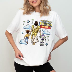 Puede incluir: Camiseta blanca con un gr&aacute;fico colorido de personajes de dibujos animados y el texto "YOU'VE GOT A FRIEND IN ME". El dise&ntilde;o incluye una figura con una t&uacute;nica amarilla, un vaquero y un guardabosques espacial, con nubes y estrellas.