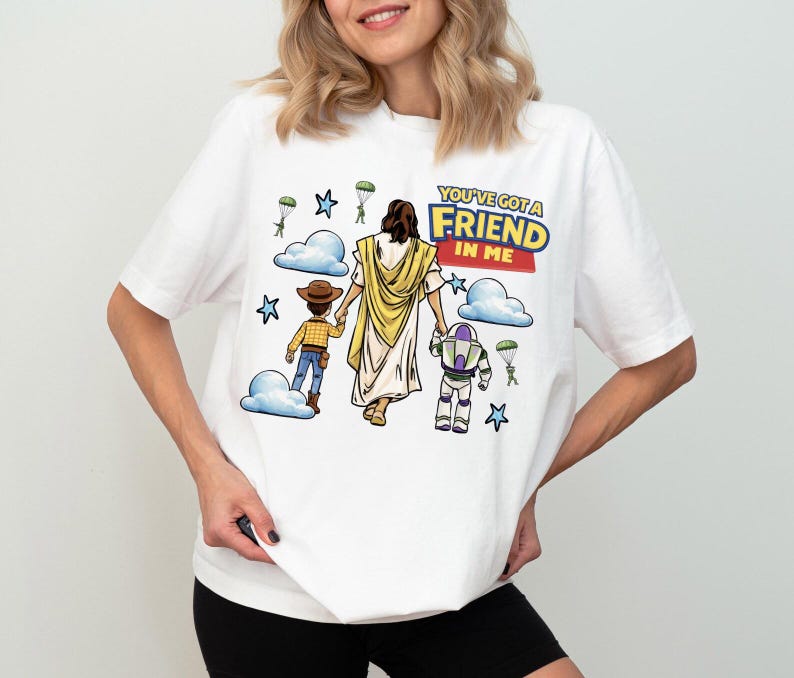 Puede incluir: Camiseta blanca con un gr&aacute;fico colorido de personajes de dibujos animados y el texto "YOU'VE GOT A FRIEND IN ME". El dise&ntilde;o incluye una figura con una t&uacute;nica amarilla, un vaquero y un guardabosques espacial, con nubes y estrellas.
