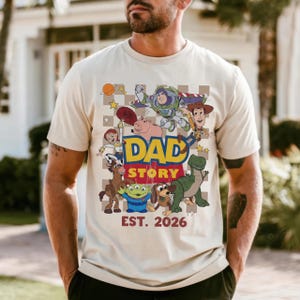 May include: Beige t-shirt featuring a colorful graphic with the text "DAD STORY EST. 2026" and various cartoon characters. The design includes a checkered background and a playful, nostalgic theme.