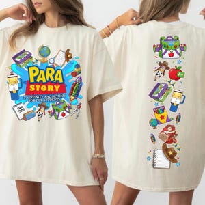 Op de afbeelding: Een off-white t-shirt met een kleurrijke grafische print. De voorkant toont de tekst "PARA STORY TO INFINITY AND BEYOND FOR OUR STUDENTS" omringd door school- en ruimtevaarticonen. De achterkant toont een verticale rangschikking van de iconen.