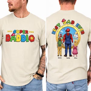 May include: A beige t-shirt featuring a graphic on the front that reads SUPER DADDIO in colourful block letters. The back displays a circular scene with a character in a red and blue outfit holding hands with two children, with text reading BEST DAD EVER.