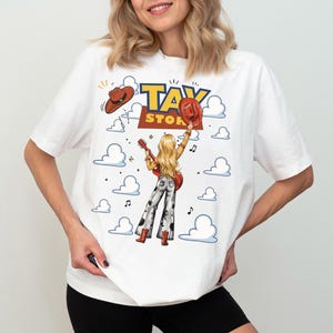 Op de afbeelding: Een wit t-shirt met een grafische print van een blond persoon met een gitaar, gekleed in een broek met wijde pijpen en rode laarzen, met een rode cowboyhoed in de hand. Het ontwerp bevat de tekst TAY STORY, zwevende wolken en muzieknoten.