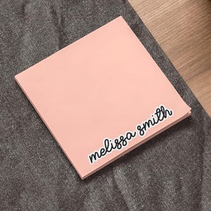 Könnte beinhalten: Ein quadratischer, hellpfirsichfarbener Haftnotizzettel mit dem Namen "melissa smith", der in einer schwarzen, kursiven Schriftart gedruckt ist. Der Zettel liegt auf einer dunkelgrauen, strukturierten Oberfläche, mit einem silbernen Stift unten links.