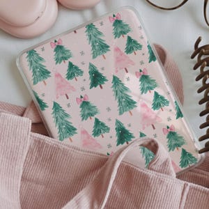 May include: A clear phone case featuring a repeating pattern of green and pink Christmas trees with bows and snowflakes on a light pink background. The case is partially visible, resting in a pink corduroy bag.