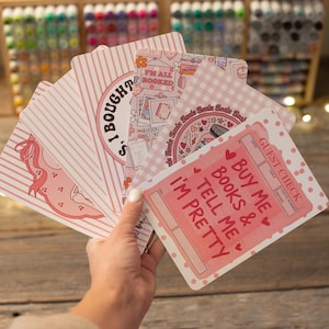 May include: A hand holds a stack of pink and white cards with various designs and text. The cards feature phrases like "I'm all booked" and "Buy me books & tell me I'm pretty". The cards have a cute, feminine aesthetic.