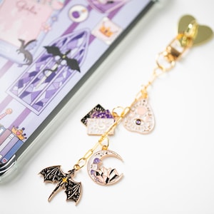 May include: A gold-colored charm bracelet with various charms, including a black dragon, a crescent moon with crystals, and a pink teardrop shape. The charms are attached to a gold chain. The background includes a phone case with a purple and white design.