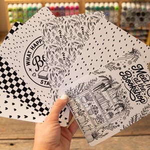 May include: A hand holds a stack of white cards with black designs. The cards feature various patterns, including polka dots, a checkerboard, and floral motifs. Some cards have text that reads "What Happ" and "Meet me at the Bookshop."