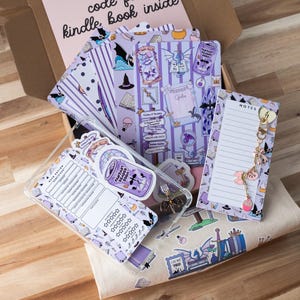 Puede incluir: Una caja llena de artículos de papelería y accesorios con temática de fantasía. Incluye cuadernos estampados, pegatinas, una funda para teléfono y un bloc de notas con un charm. Los artículos presentan ilustraciones de dragones, murciélagos y otros elementos mágicos en tonos morados y blancos.