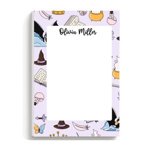 May include: A lavender notepad with a white interior and a border featuring whimsical illustrations of witch hats, books, cauldrons, and butterflies. The name "Olivia Miller" is written in elegant script.