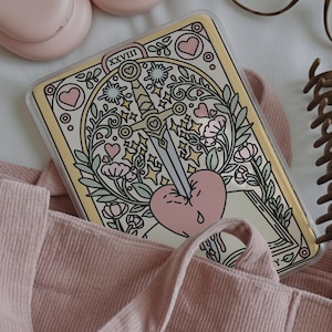 May include: A clear phone case featuring a tarot card design. The card depicts a sword piercing a pink heart, surrounded by floral and star motifs. The case has a cream-colored border and is set against a pink background.