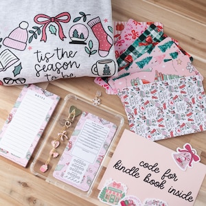Puede incluir: Una colección de artículos de papelería y accesorios con temática navideña. Incluye una sudadera gris con un gráfico festivo, papel estampado, una funda transparente con un dije, un bloc de notas y pegatinas. El texto "tis the season to be" es visible.