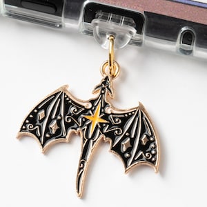 May include: A gold-colored dragon charm with black enamel wings and body, and a yellow starburst design. The charm is attached to a clear phone case with a gold-colored ring.