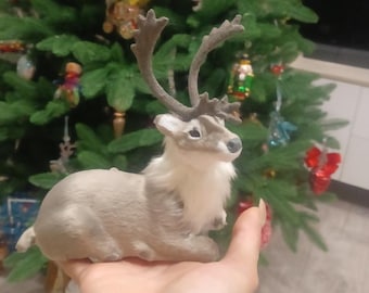 May include: A plush gray reindeer figurine with brown antlers and a white fur trim. The reindeer is lying down with its head turned to the side. The figurine is being held in a person's hand.