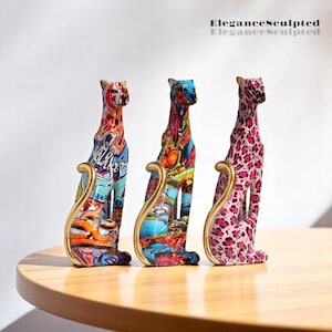 Peut inclure: Trois sculptures de chats en céramique colorées avec des accents dorés. Les chats sont assis et présentent différents motifs, dont un imprimé léopard rose, un motif graffiti bleu et orange et un motif graffiti rouge et vert.