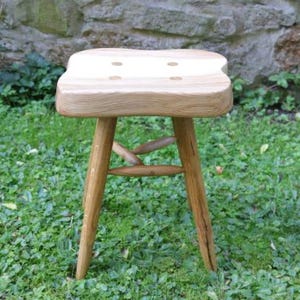 Puede incluir: Un taburete de madera con un asiento rectangular de color claro con esquinas redondeadas y cuatro incrustaciones circulares. El taburete tiene tres patas cónicas conectadas por una estructura de soporte central de madera, mostrando un acabado de veta natural.