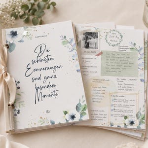 May include: A guestbook with a white cover featuring blue and green floral illustrations, gold speckles, and the German text "Die schönsten Erinnerungen sind ganz besondere Momente". The book is bound with a beige satin ribbon on the left side.
