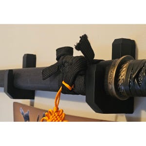 May include: A black katana sword rests horizontally on a black wall-mounted display stand. The scabbard features a black cord wrap with a bright orange decorative tassel hanging from the center. A metallic tsuba guard sits near the handle.