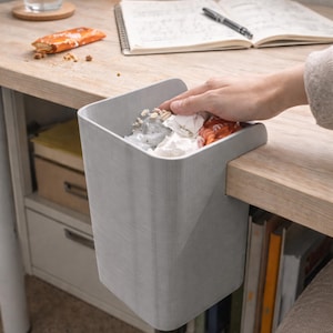 Puede incluir: Un cubo de basura de escritorio gris claro está sujeto al borde de un escritorio de madera. El cubo está lleno de papel arrugado y envoltorios de comida. Una mano está metiendo la mano en el cubo. Un cuaderno, un bolígrafo y un tentempié están en el escritorio.