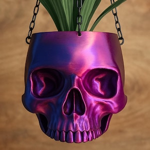 May include: A hanging planter shaped like a human skull with a metallic iridescent finish that shifts between shades of purple, magenta, and blue. The skull features deep eye sockets and a nasal cavity, suspended by three black metal chains.