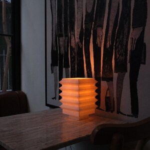 Op de afbeelding: Een verlichte, vierkante papieren lantaarn met een zigzagrand staat op een houten tafel. Het warme licht van de lantaarn verlicht de tafel en het kunstwerk aan de muur erachter. Het kunstwerk is een zwart-wit schilderij van figuren.