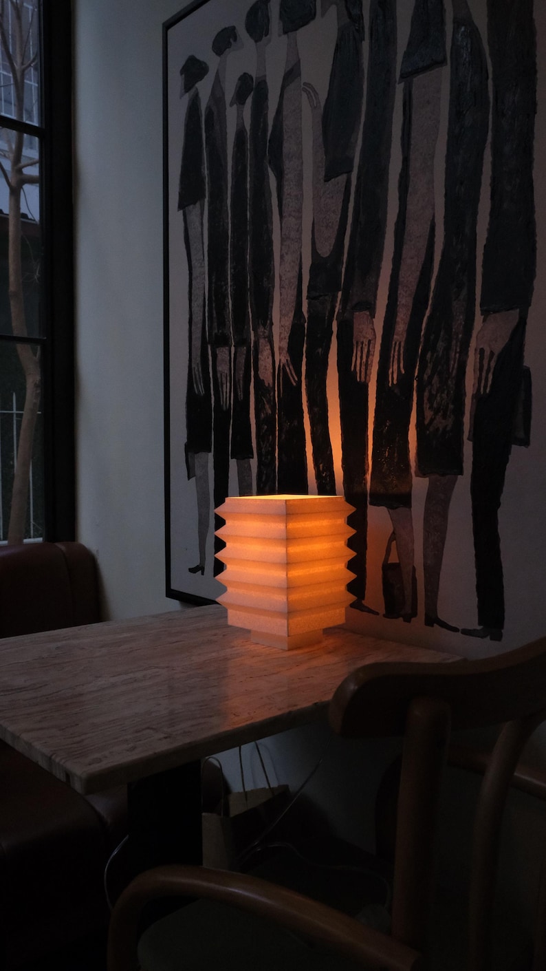 Op de afbeelding: Een verlichte, vierkante papieren lantaarn met een zigzagrand staat op een houten tafel. Het warme licht van de lantaarn verlicht de tafel en het kunstwerk aan de muur erachter. Het kunstwerk is een zwart-wit schilderij van figuren.