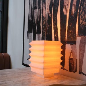 Op de afbeelding: Een wit en gele, geometrisch gevormde lamp met een warme gloed staat op een lichtgekleurde tafel. De lamp heeft een gelaagd ontwerp met een zigzagrand. Op de achtergrond een schilderij met figuren in zwart en wit.