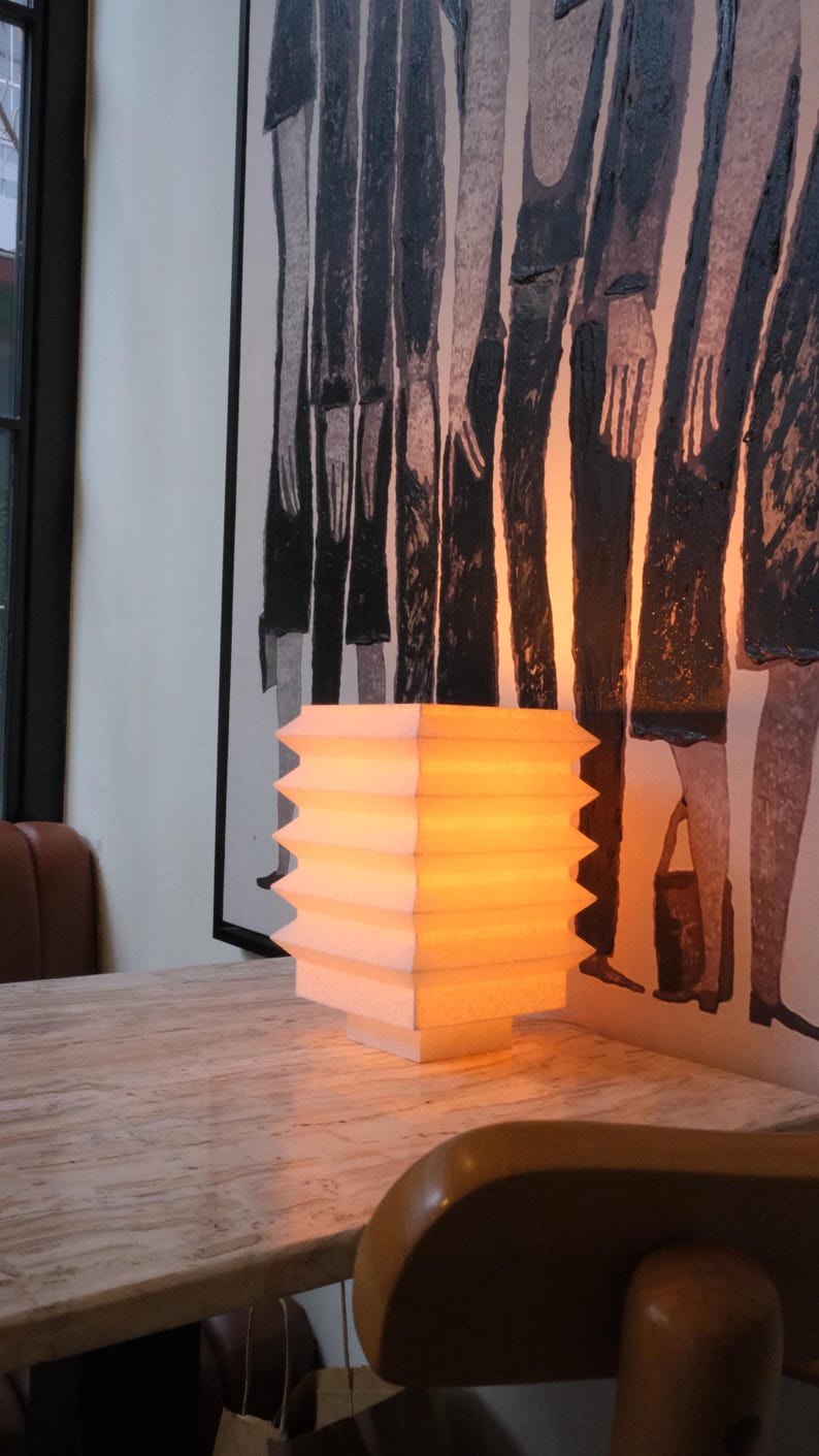 Op de afbeelding: Een wit en gele, geometrisch gevormde lamp met een warme gloed staat op een lichtgekleurde tafel. De lamp heeft een gelaagd ontwerp met een zigzagrand. Op de achtergrond een schilderij met figuren in zwart en wit.