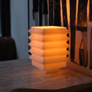 Op de afbeelding: Een rechthoekige lamp met een gelaagd ontwerp, die een warm, geel licht uitstraalt. De lamp is gemaakt van een lichtgekleurd materiaal en staat op een tafel met marmerpatroon. De achtergrond toont een abstract kunstwerk in zwart en wit.