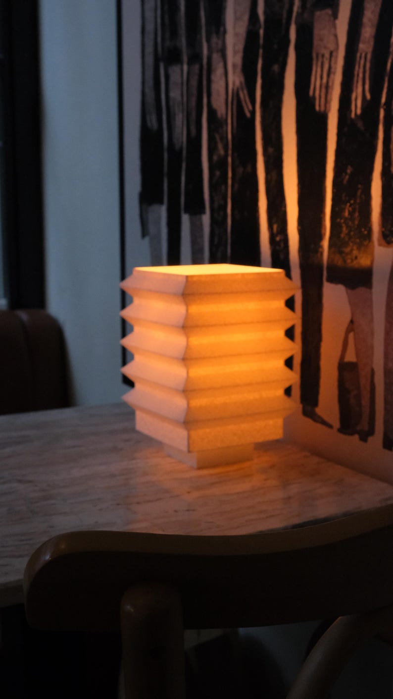 Op de afbeelding: Een rechthoekige lamp met een gelaagd ontwerp, die een warm, geel licht uitstraalt. De lamp is gemaakt van een lichtgekleurd materiaal en staat op een tafel met marmerpatroon. De achtergrond toont een abstract kunstwerk in zwart en wit.