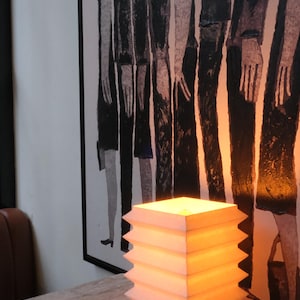 Op de afbeelding: Een witte, geometrische lamp met een warm, gloeiend licht staat op een marmerpatroon oppervlak. Het ontwerp van de lamp heeft een reeks gestapelde, zigzagvormige lagen. Een zwart-wit kunstwerk staat op de achtergrond.