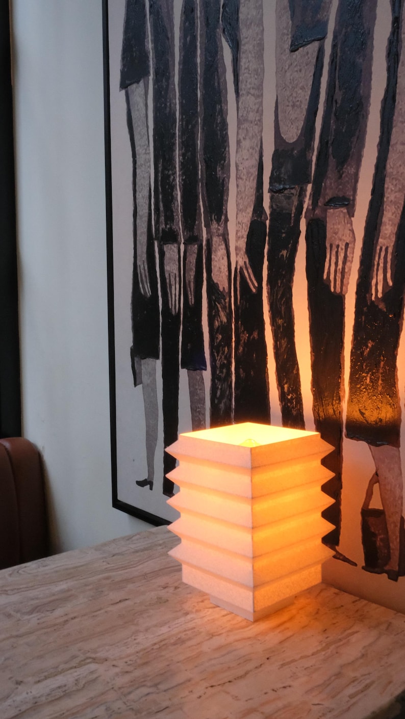 Op de afbeelding: Een witte, geometrische lamp met een warm, gloeiend licht staat op een marmerpatroon oppervlak. Het ontwerp van de lamp heeft een reeks gestapelde, zigzagvormige lagen. Een zwart-wit kunstwerk staat op de achtergrond.