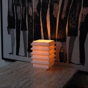 Op de afbeelding: Een kleine, witte accordeonlamp straalt een warme gloed uit en staat op een tafel met marmerlook. Het ontwerp van de lamp heeft een reeks gelaagde zigzagranden. Op de achtergrond hangt een zwart-wit kunstwerk.