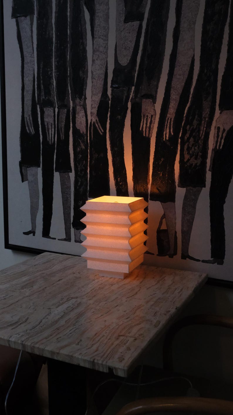 Op de afbeelding: Een kleine, witte accordeonlamp straalt een warme gloed uit en staat op een tafel met marmerlook. Het ontwerp van de lamp heeft een reeks gelaagde zigzagranden. Op de achtergrond hangt een zwart-wit kunstwerk.