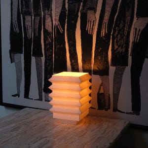 Op de afbeelding: Een moderne tafellamp met een geometrisch ontwerp. De lamp heeft een gestapeld, harmonica-achtig ontwerp in een lichtbeige kleur en straalt warm licht uit. Hij staat op een getextureerde tafel, met een abstract zwart-wit kunstwerk op de achtergrond.