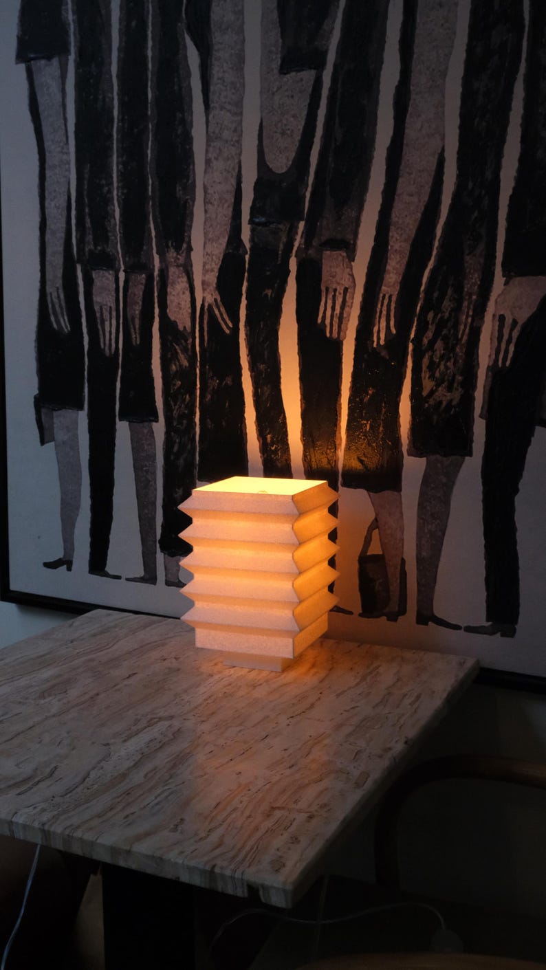 Op de afbeelding: Een moderne tafellamp met een geometrisch ontwerp. De lamp heeft een gestapeld, harmonica-achtig ontwerp in een lichtbeige kleur en straalt warm licht uit. Hij staat op een getextureerde tafel, met een abstract zwart-wit kunstwerk op de achtergrond.