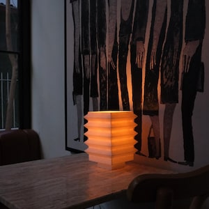 Op de afbeelding: Een verlichte, vierkante papieren lantaarn met een zigzagrand staat op een marmeren tafel. Het warme licht van de lantaarn verlicht de tafel en de omgeving. Een zwart-wit schilderij hangt aan de muur.