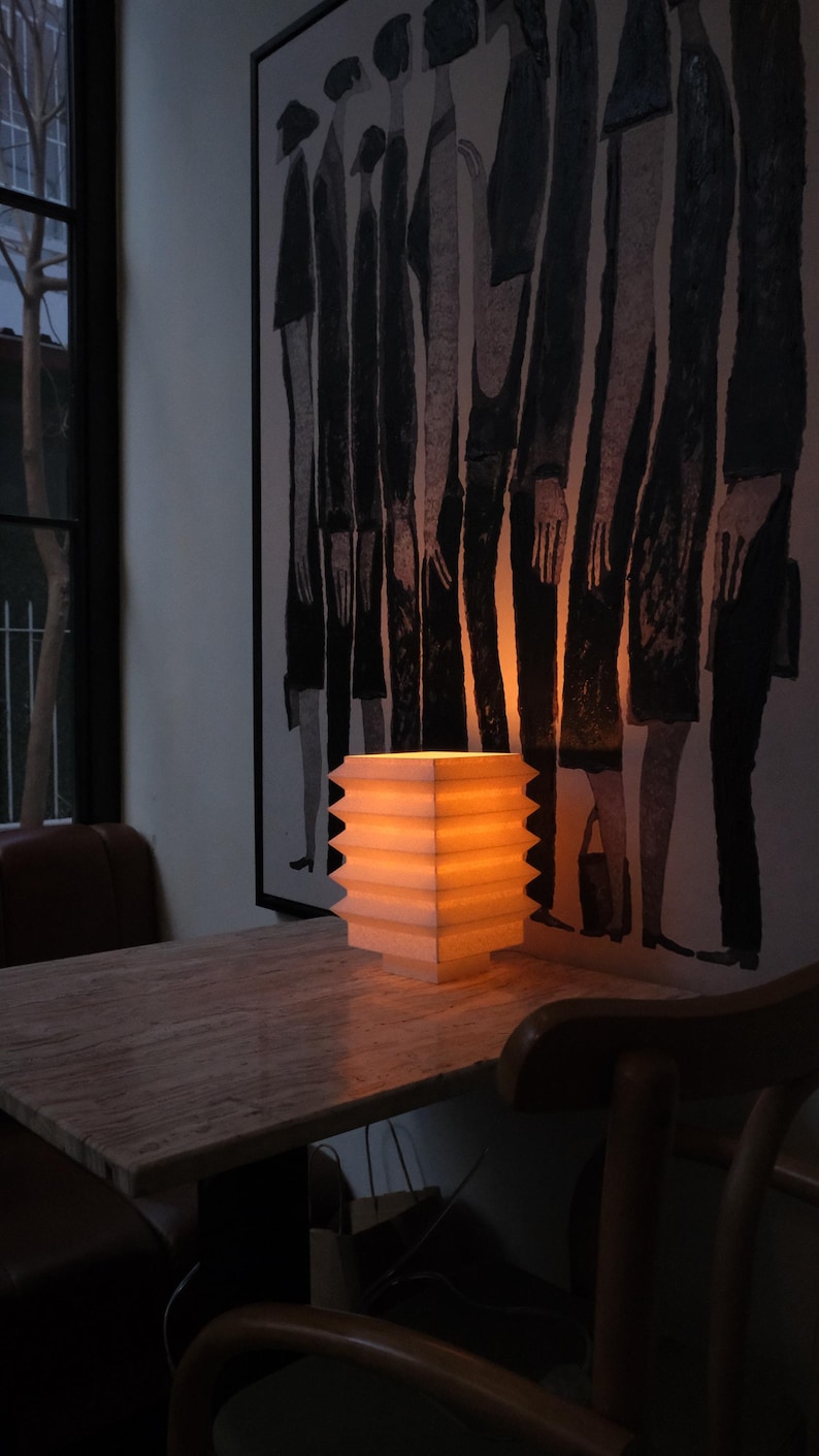 Op de afbeelding: Een verlichte, vierkante papieren lantaarn met een zigzagrand staat op een marmeren tafel. Het warme licht van de lantaarn verlicht de tafel en de omgeving. Een zwart-wit schilderij hangt aan de muur.