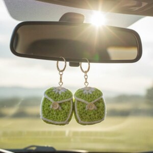 Op de afbeelding: Twee groen-witte gehaakte auto-hangers hangen aan een achteruitkijkspiegel. De hangers zijn vierkant met knoopdetails en een witte rand. De achtergrond toont een zonnige buitensc&egrave;ne.